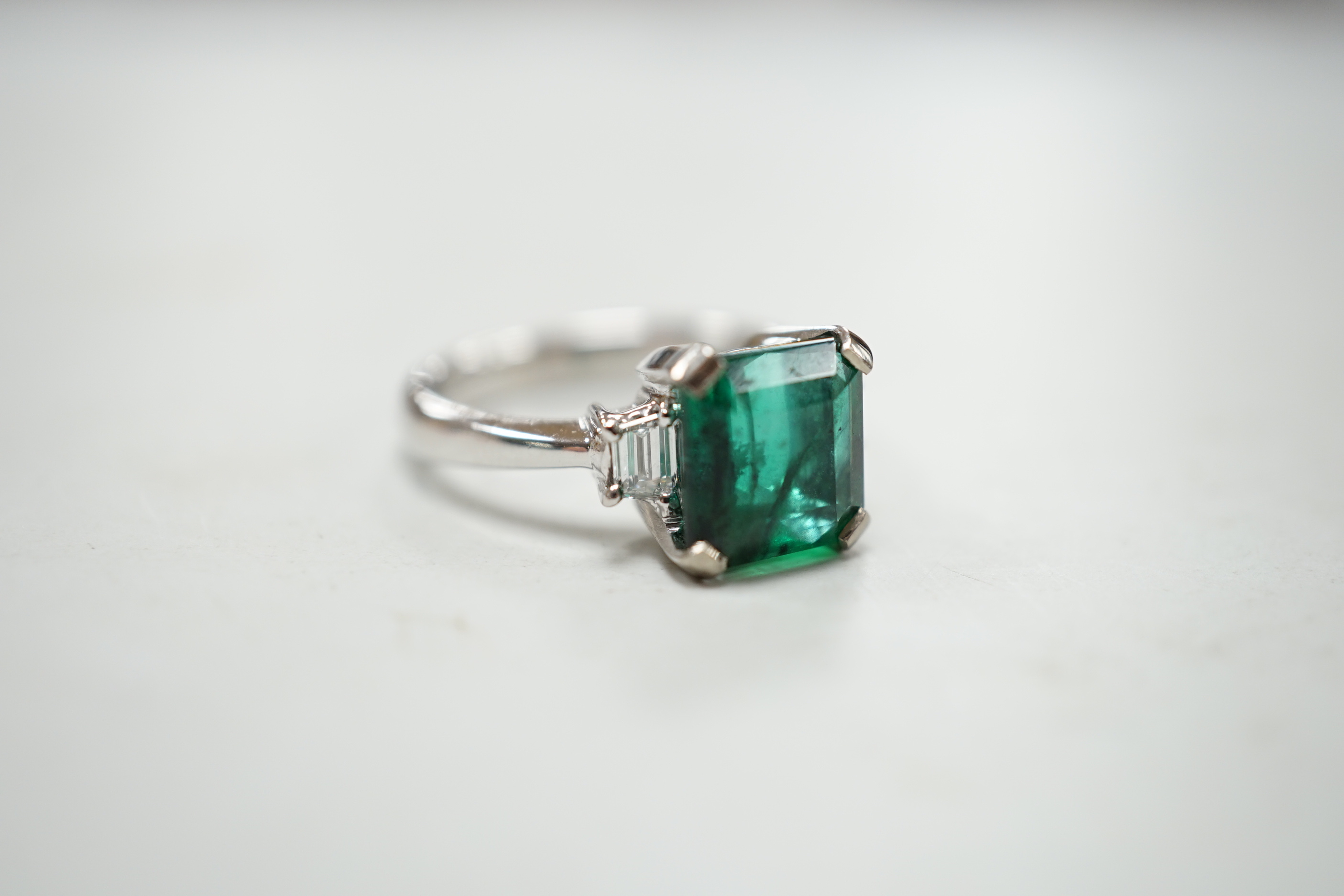 A modern 18k white metal and single stone emerald set ring, baguette cut diamond set shoulders, size N, gross weight 5.8 grams